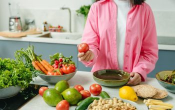 Woman in pink overshirt holds tomato in kitchen with fresh fruits and veggies.